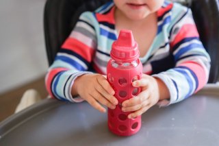 Test der neuen Kaffetasse für Piloten. Little girl in high-chair holds glass sippy cup with rubber seal and hard spout with water